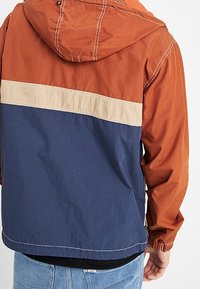 Color-block jas met een oranje capuchon, een crème horizontale streep en een marineblauwe romp. Gemaakt van lichtgewicht stof met elastische manchetten.