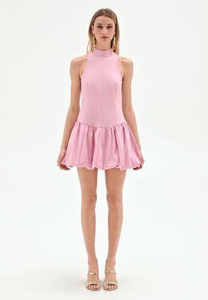 Femme portant une robe rose sans manches à col montant, avec une jupe froncée et évasée, et des talons ouverts dorés, debout sur un fond blanc.