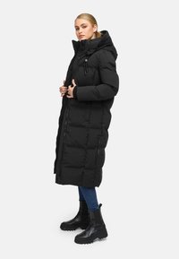 Black long puffer coat with a hood, featuring quilted texture, zip closure, and side pockets. Worn with blue jeans and black ankle boots.