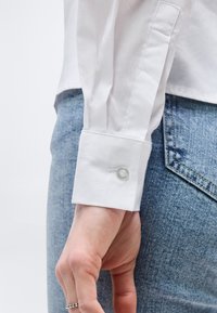 Χέρι με ασημένιο δαχτυλίδι με χάντρες δίπλα σε μανσέτα λευκού πουκαμίσου με κουμπί, ακουμπισμένο κοντά στην εμπρόσθια τσέπη ανοιχτού μπλε τζιν παντελονιού.