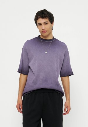 Fiatal férfi rövid sötét hajjal, lila rövid ujjú inget és fekete nadrágot visel, egyszerű fehér háttér előtt áll.