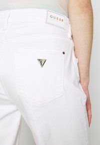 Jeans di denim bianchi con un'etichetta triangolare dorata sulla tasca posteriore. Caratteristiche includono un design standard a cinque tasche e cuciture visibili.