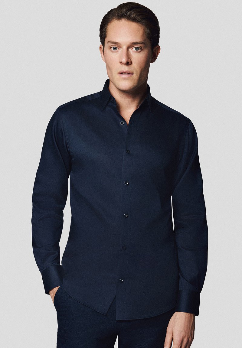 Granatowa koszula z długim rękawem, zapinana na guziki, o gładkiej fakturze, z wywiniętym kołnierzykiem i czarnymi guzikami. Posiada dopasowany krój oraz prosty dół.