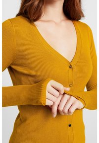 Senapsgul ribbad cardigan med V-hals, långa ärmar och knappdetaljer fram. Den mjuka texturen betonar värme och komfort.
