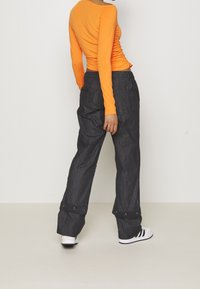 Orange långärmad topp med rynkning på sidan; matchad med lösa, mörka denimbyxor med tryckknappar vid bensluten och sidofickor. Vita sneakers synliga.
