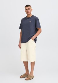 Camiseta de algodón azul oscuro con un emblema del sol en el pecho, combinada con pantalones cortos crema claros y sandalias marrones. Corte recto, diseño casual.