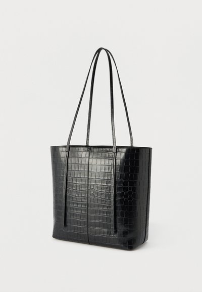 Μαύρη τσάντα tote με υφή κροκοδείλου, δύο μακριές λαβές, ορθογώνιο σχήμα και λεία επιφάνεια σε ομοιόμορφο ανοιχτό φόντο.