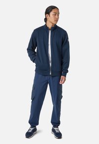 Giacca a zip blu in tessuto morbido, abbinata a pantaloni cargo blu con tasche laterali. Il modello indossa scarpe da ginnastica nere e grigie.