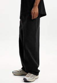 Person som bär lösa svarta byxor och beige sneakers som står mot en vit bakgrund.