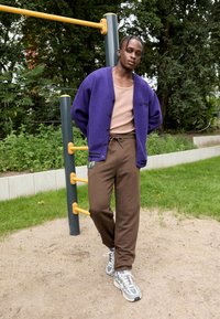 Cardigan oversize viola, camicia fitted color beige, pantaloni della tuta marroni con coulisse e scarpe da ginnastica grigie. Ambientazione all'aperto con vegetazione.