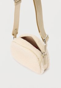 Pequeño bolso cruzado de pelaje color beige con correa de tela ajustable y cremallera parcialmente abierta que muestra el interior color beige.