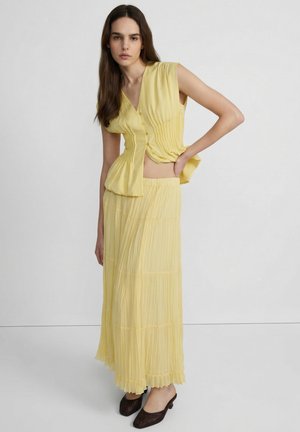 Kobieta z długimi brązowymi włosami, nosząca żółty top bez rękawów z guzikami i pasującą plisowaną maxi spódnicę, stojąca z jedną ręką na biodrze.