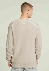 Maglione a costine beige con collo a giro, maniche raglan e una piccola etichetta sulla parte superiore della schiena, indossato sopra jeans in denim chiaro.