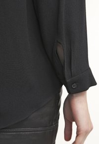 Blouse noire en tissu texturé, dotée d'un col plongeant et d'un poignet boutonné avec une fente latérale près du poignet. Gros plan sur la manche.