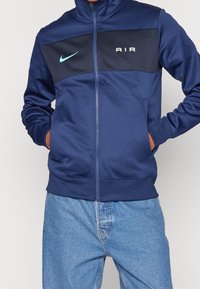 Mörkblå zip-up jacka i slätt tyg, med en mörkblå panel som har ordet "AIR" och en ljusblå Nike-logotyp på bröstet.