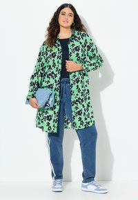 Camicia oversize con stampa leopardata verde e nera, abbinata a jeans blu scuro e sneakers azzurro chiaro, mentre tiene una borsa azzurro chiaro.