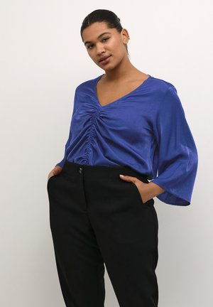 Blaue Satinbluse mit V-Ausschnitt und gerafftem Mittelteil. Mit langen, ausgestellt geschnittenen Ärmeln und glatter Textur. Kombiniert mit schwarzen Hosen.