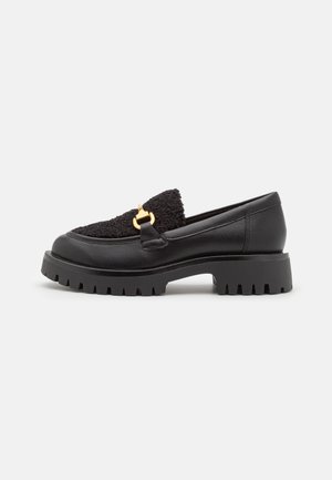 Sorte loafers med et tekstureret, fuzzy stofoverdel, der har et guldfarvet hardware-detalje og en chunky, rillet ydersål. Glat læderdetaljering på siderne.