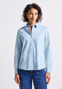 Vrouw met krullend haar, draagt zilveren oorbellen, een lichtblauwe verticaal gestreepte blouse en blauwe spijkerbroek tegen een neutrale achtergrond.