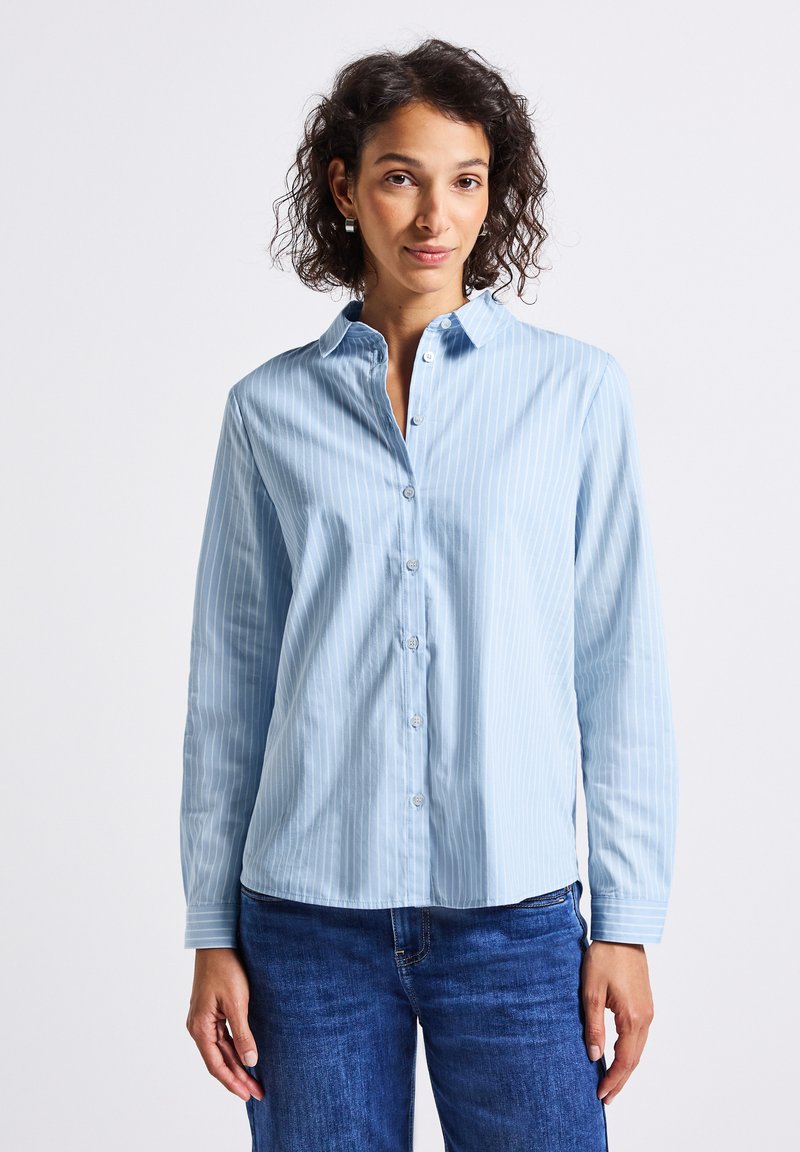 Vrouw met krullend haar, draagt zilveren oorbellen, een lichtblauwe verticaal gestreepte blouse en blauwe spijkerbroek tegen een neutrale achtergrond.