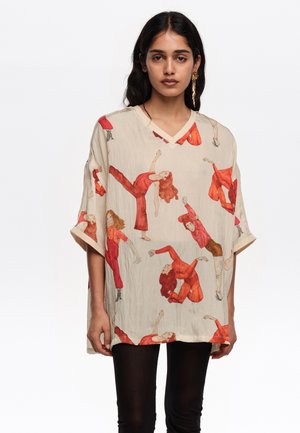 Mujer con camisa beige de cuello en V de gran tamaño que presenta ilustraciones de bailarines en rojo y naranja, combinada con pantalones negros y un largo pendiente dorado.