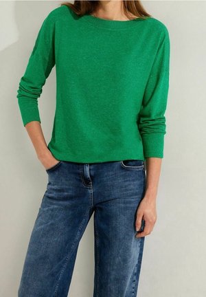 Femme portant un pull vert vif à manches longues avec col rond et un jean bleu, debout avec une main dans la poche sur un fond uni.