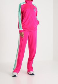 Persona vestida con un chándal rosa brillante con rayas azul claro y blancas en las mangas y los pantalones, combinado con zapatillas blancas, de pie.