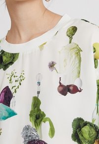 Weißes Oberteil aus Stoff mit buntem Gemüse-Print, darunter Salat, Zwiebeln, Kräuter und Glühbirnen, mit einem gerippten Halsauschnitt.