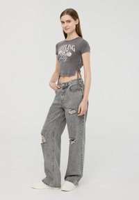 T-shirt grigio corto con grafica "Feeling Lucky", abbinato a jeans larghi strappati di un grigio chiaro. La modella indossa scarpe da ginnastica bianche.