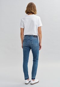 Jean skinny bleu en denim avec taille mi-haute, comprenant deux poches arrière et un patch en cuir noir. Associé à un T-shirt blanc à manches courtes.