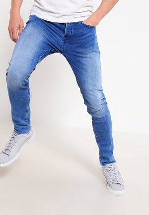 Jean en denim bleu ajusté avec des détails décolorés, doté de cinq poches et d'une fermeture éclair, associé à des baskets grises claires.