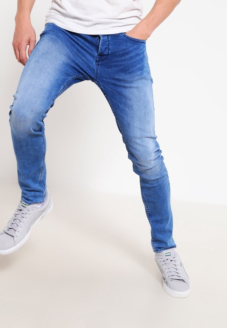 Jeans de denim azul ajustados con detalles de desgaste, que cuentan con cinco bolsillos y cierre de cremallera, combinados con zapatillas gris claro.