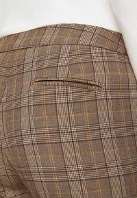 Pantalon à carreaux marron en tissu texturé, avec une poche arrière et de fines rayures jaunes entrelacées de carreaux plus foncés.