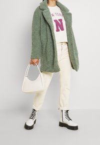 Grön fuskpälsjacka med notch-krage, krämfärgad sweatshirt med "New York"-tryck, beige kordflöjtsbyxor och vita snörstövlar. Vit handväska.