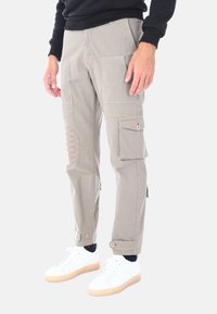 Pantaloni cargo beige con molte tasche, caratterizzati da cuciture decorative rosse nella zona del ginocchio e polsini fissati. Indossati con sneaker bianche.