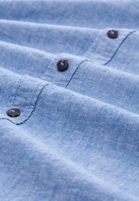 Lichtblauw shirtstof met een gestructureerde uitstraling, voorzien van donkere knopen. De stof is zacht en licht kreukelig met zichtbare stikdetails.
