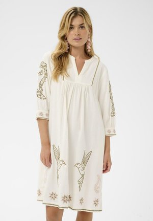 Ženska, oblečena v belo obleko dolžine do kolen s tremi četrtinami rokavov, vezenimi pticami in cvetličnimi vzorci ter roza uhani z resicami.