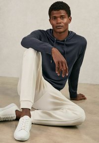 Hanorac cu glugă de culoare bleu marin, pantaloni joggeri crem cu croială conică și adidași albi. Materiale moi, croială lejeră, cu șnur la gât, design minimalist.