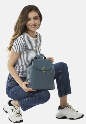 Sac à dos en simili cuir bleu avec un haut arrondi, quincaillerie dorée, fermeture à boucle à l'avant et fermeture éclair sur le côté. Le modèle pose en tenant le sac.