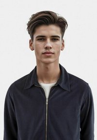 Giacca blu navy con zip, dotata di colletto a punta e realizzata in tessuto testurizzato, indossata sopra una maglietta bianca, con una zip metallica ben visibile.