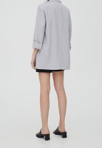 Personne debout de dos, portant un blazer gris clair, un short noir et des sandales à talons blocs noires à bout ouvert sur un fond uni.