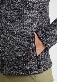 Giacca in tessuto nero e grigio melangiato con tasca laterale dotata di una cerniera nera e un piccolo dettaglio arancione vicino all'orlo. Design a maniche lunghe con texture.
