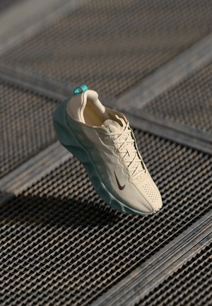 Beige athletic shoe with teal accents, textured upper, and brown Nike swoosh. Features a rounded toe and cushioned sole. Floating above a patterned surface.