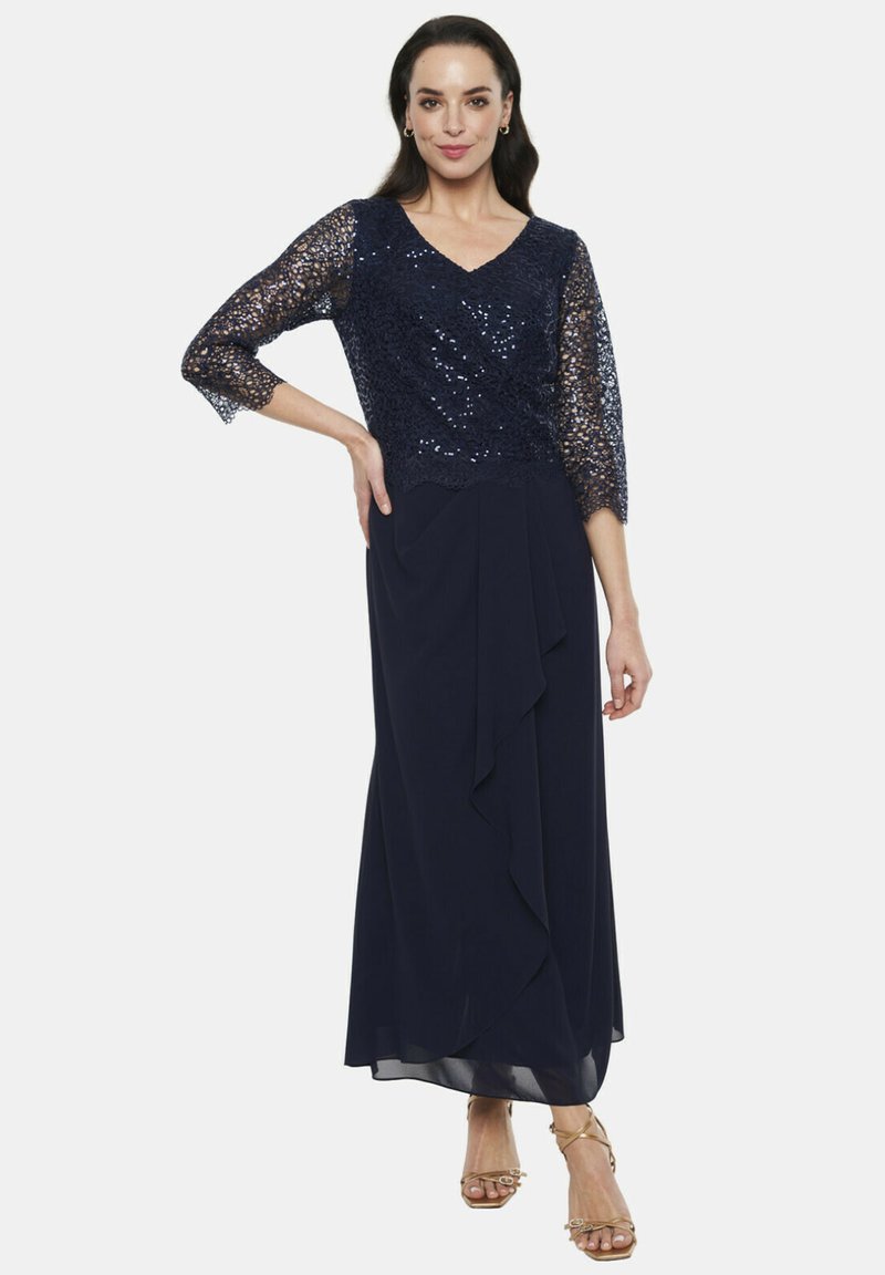 Długa granatowa sukienka z koronkową górą ozdobioną cekinami i rękawami o długości trzech czwartych. Zwiewna spódnica z drapowanym detalem. W połączeniu z sandałami na cienkich paskach w kolorze złotym.