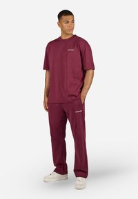 Bordeauxfarbenes Set aus T-Shirt und Hose aus Baumwolle. Das T-Shirt hat einen Rundhalsausschnitt und kurze Ärmel; die Hose verfügt über Seitentaschen und Branding.