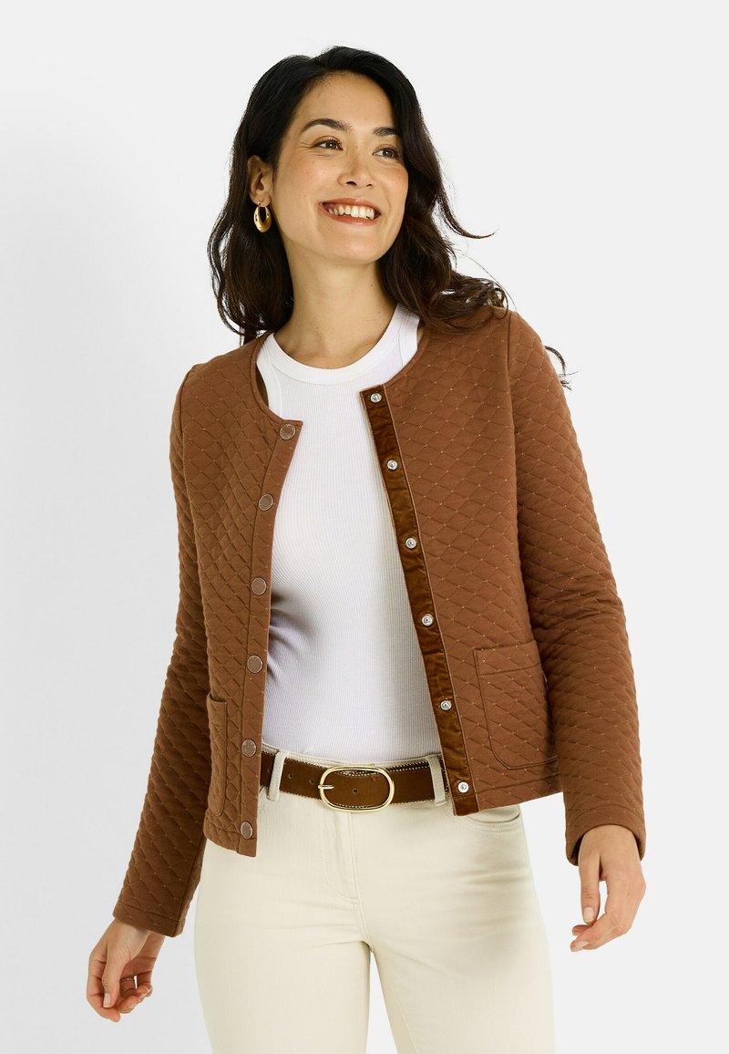 Chaqueta marrón acolchada con cierre de botones, escote redondo y dos bolsillos delanteros, combinada con una camiseta blanca y pantalones de color crema.