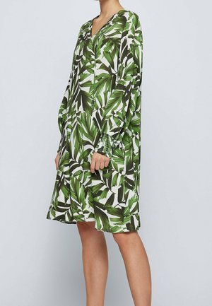 Femme portant une robe longue jusqu'aux genoux, à manches longues, avec un imprimé de feuilles tropicales vertes, un col en V et des poignets froncés sur un fond clair uni.