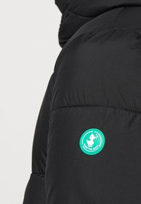 Sort pufferjakke med et rundt grønt logo-patch med en and-illustration på skulderen. Materialet ser glat og puffy ud.