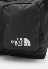Mochila de tecido preta com um grande logotipo branco da The North Face. O design inclui um bolso com fecho e uma superfície texturizada.