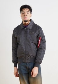 Dark gray bomber jacket with front zipper, ribbed cuffs, and a patch on the chest. Features a red tag on the sleeve. Worn with blue jeans.